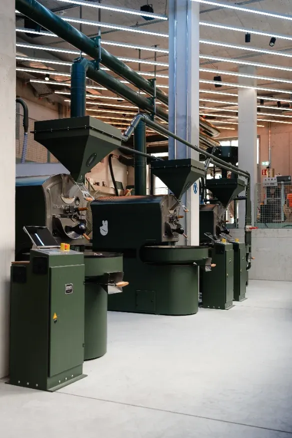 Line of dark-green roasting machines with hoppers and ducting in the Munich roast hall